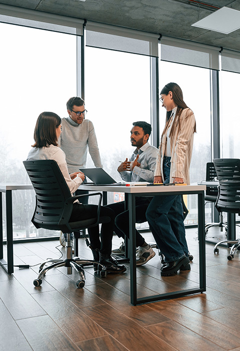 four professionals collaborating around a laptop in a modern office space discussing innovative ideas while seated at a table as part of a team meeting focused on strategy and solutions for three key projects