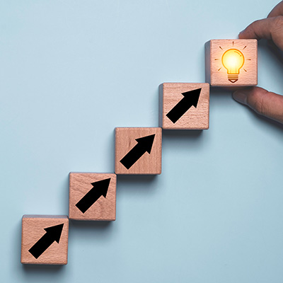 hand holding wooden block steps with upward arrows and a glowing light bulb representing growth and progress through six steps