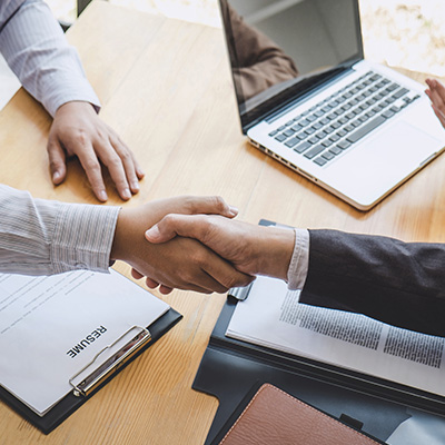 two people shaking hands during a meeting with resumes on the table and a laptop visible in the background